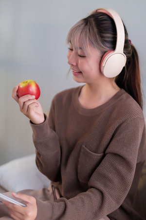 Capture a young Asian woman with headphones relishing a fresh apple indoors depicting wellness relaxation and healthy lifestyle choice for radiant skin and balanced diet.の写真素材