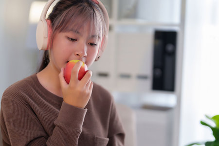 Young Asian woman is listening to music with headphones and eating a red apple, a healthy snack for lifestyle and wellbeing while relaxing at home.の写真素材