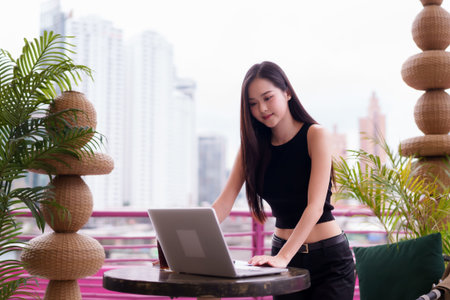 Confident Asian adult woman works remotely on laptop from a stylish urban balcony. Embracing digital nomad lifestyle with city view.の写真素材