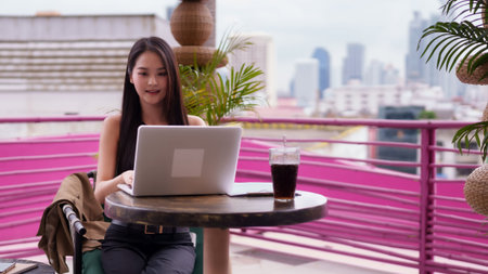 Confident Asian adult woman works remotely on laptop at vibrant rooftop cafe. Urban skyline provides modern backdrop for digital nomad lifestyle.の写真素材