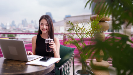 Confident Asian adult woman uses a laptop computer for remote work outdoors on a vibrant city balcony She embodies modern productivity and urban lifestyleの写真素材
