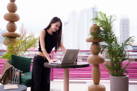Confident Asian adult woman uses a laptop on an urban balcony She is focused on remote work in a modern city environmentの写真素材