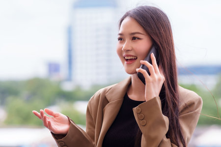 Modern Asian businesswoman engages in a vibrant smartphone conversation outdoors Her expressive gesture conveys dynamic communication and urban connectivityの写真素材