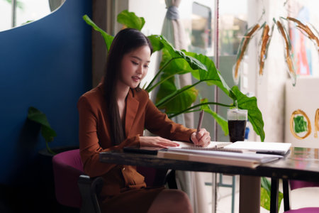 Confident Asian woman focused on professional tasks in a contemporary cafe setting. Her dedicated work ethic creates a productive atmosphere.の写真素材