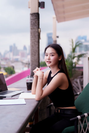 Confident Asian woman working remotely in chic rooftop setting with city view. Professional female freelancer embracing digital nomad lifestyle.の写真素材