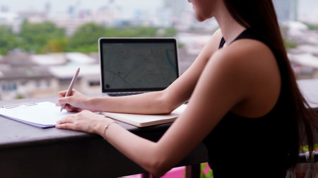 Focused Asian woman works remotely from a modern balcony office. She meticulously writes notes on a clipboard while using her laptop. This professional individual demonstrates dedicated productivity in an inspiring urban setting.の写真素材