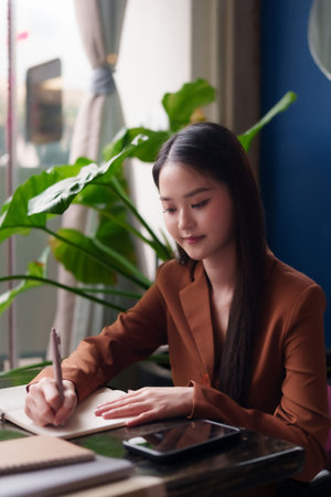 A focused asian adult woman writes notes in a journal. She is a dedicated professional succeeding in her modern work environment.の写真素材