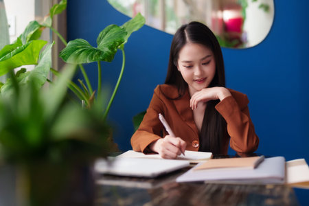 Confident Asian professional diligently taking notes at her desk in a stylish office environment. Empowered woman focused on her modern work.の写真素材