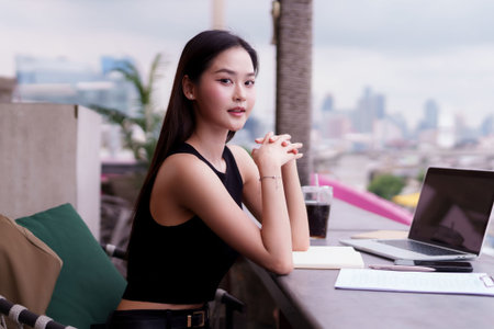 Confident Asian adult woman works smart on laptop at a chic rooftop cafe. She plans her career...