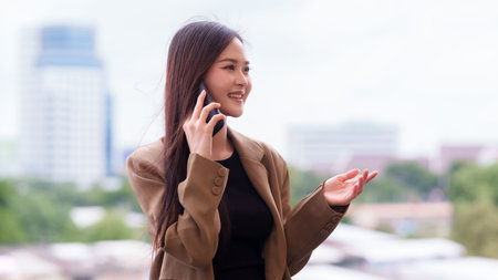 Adult Asian professional female uses smartphone outdoors. She smiles discussing business confident communication lifestyle modern urban setting.の写真素材