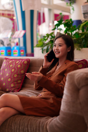 Smiling Asian adult woman talks on smartphone in a modern lounge. She radiates confidence and professionalism while engaging in digital communication.の写真素材