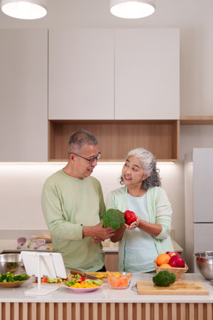 Happy senior Asian man and woman cook together in a modern kitchen. They hold fresh vegetables for a nutritious dish.の写真素材
