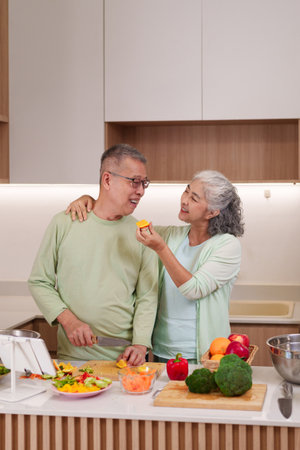 Asian older couple enjoy preparing healthy meal Woman feeds man orange slice Man chops vegetables Happy bonding kitchen activity Fresh ingredientsの写真素材