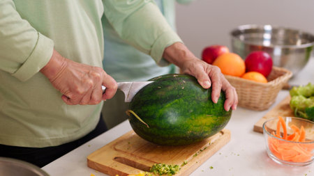 Closeup shot adult hands carefully slicing ripe watermelon with sharp knife on wooden board kitchen counter surrounded by colorful fruits vegetables food prepの写真素材