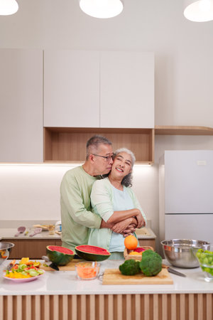 Happy Asian elderly couple embracing in a modern kitchen with fresh fruits and vegetables preparing a nutritious meal enjoying domestic life.の写真素材