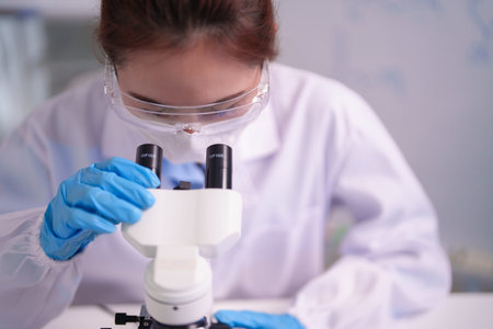 Focused Asian adult woman scientist observes sample through microscope in laboratory. Wearing lab...