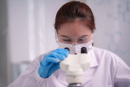 Adult Asian female scientist in lab coat goggles mask gloves intently examining sample through microscope laboratory environmentの写真素材
