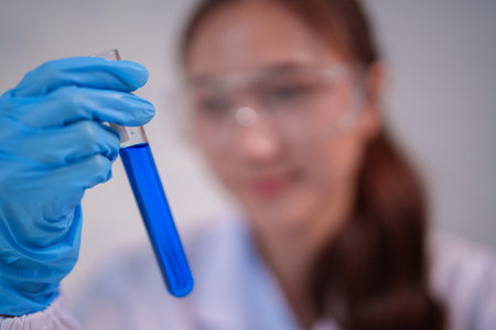 Adult Asian woman researcher conducts scientific experiment holding test tube blue liquid. Focus on precise chemical analysis and laboratory studyの写真素材