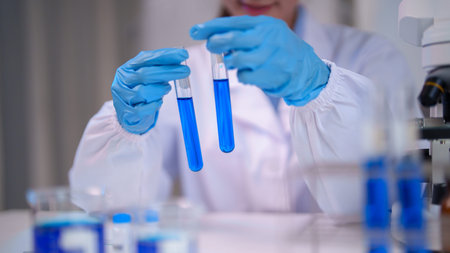 Adult Asian scientist examines blue liquid in test tubes with gloved hands performing scientific...