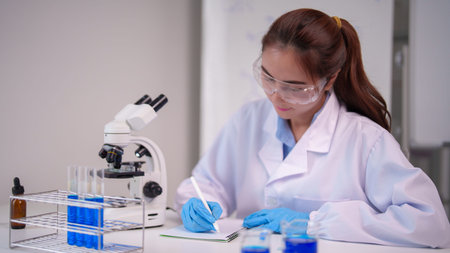 Focused Asian adult woman scientist conducts laboratory analysis. She wears protective gear, studies samples, records observations using lab equipment. Innovation research development.の写真素材