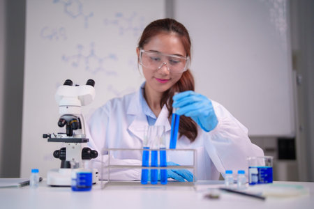 Adult Asian woman scientist in lab coat safety goggles gloves performs experiment with blue liquid...
