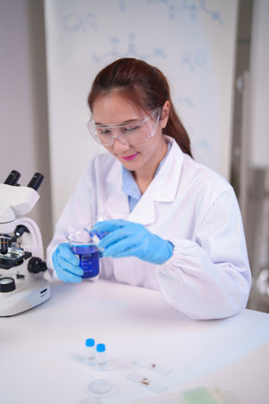 Asian adult female scientist conducts chemistry experiment pouring blue solution in modern laboratory. She wears protective gearの写真素材