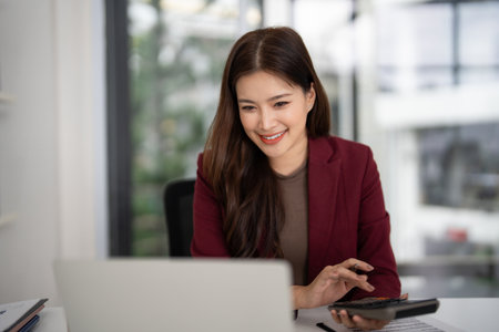 Professional Asian woman calculates real estate figures using laptop in bright office She manages property sales finances with a confident smileの写真素材