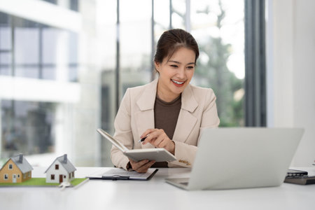 Asian adult real estate professional smiles working at modern office desk with house models and laptop managing property salesの写真素材