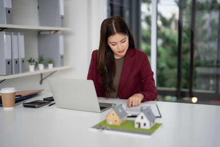Professional Asian woman agent diligently examines residential property contracts using laptop preparing for housing market investments and customer serviceの写真素材
