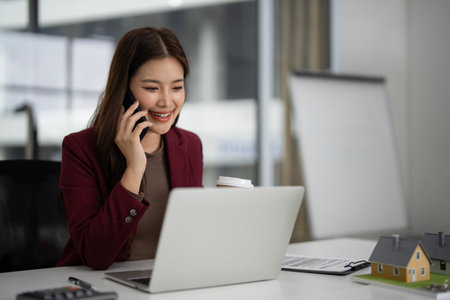 Asian professional communicates clients about property and uses laptop phone for home sales business. Modern office workspace.の写真素材