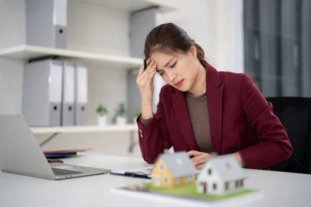 Asian adult female property agent stressed at office desk managing real estate market challenges with house models financial pressure business issuesの写真素材