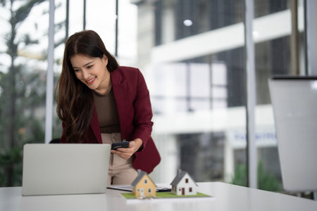 Professional Asian woman realtor smiling uses smartphone with laptop and miniature house models for property business dealsの写真素材