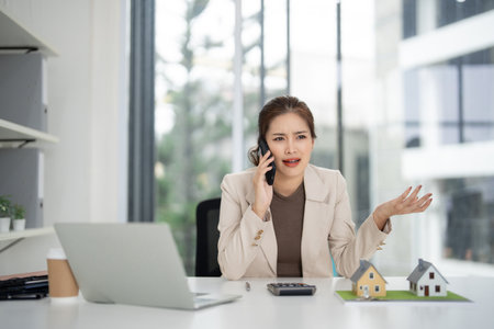 Asian adult woman realtor speaks mobile phone in a modern office. She discusses property sales using house models on her desk. Serious business communication.の写真素材