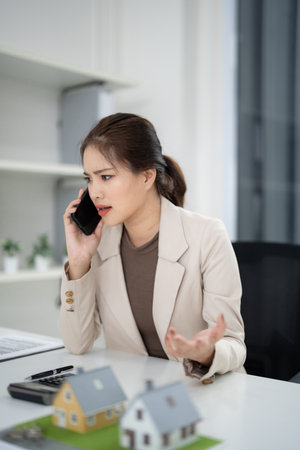 Adult Asian real estate agent has a serious phone discussion. Property models on her desk. She handles a challenging client.の写真素材