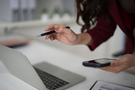 Diligent adult Asian hands engage modern technology for property sales. Multitasking professional uses laptop phone pen efficiently in office. Background blur emphasizes focus.の写真素材