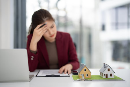 Asian adult real estate professional feels overwhelmed contemplating property market issues in a contemporary office setting with house models.の写真素材