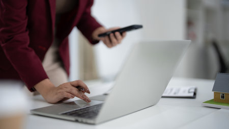Adult Asian woman professionally working. Efficient real estate agent utilizes laptop and smartphone. Manages property sales and client communications from office desk.の写真素材