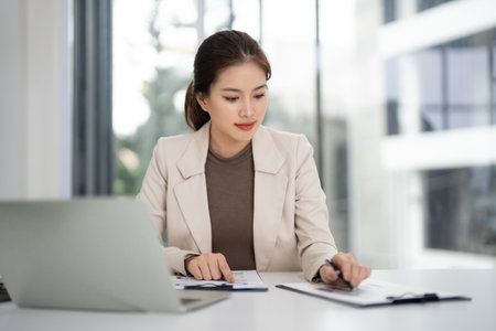 Adult Asian professional works diligently at modern office desk. She reviews important property documents for real estate sales. Laptop visible. Focused business success.の写真素材