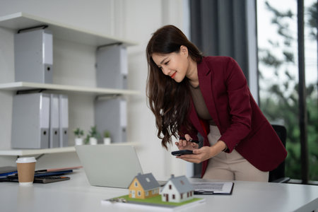 Professional Asian woman realtor reviews property details using phone. Models and laptop aid strategic house selling plans.の写真素材