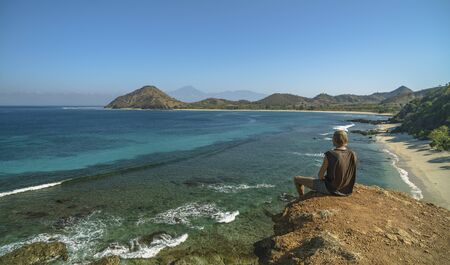 Blond man sitting and enjoying the views to undiscovered beach, hills and Rinjani Volcano seen in Sumbawa islandの写真素材