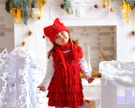 Joyful girl in red dress. Happy girl in a Christmas setting. White fireplace, white house and the white tree.のeditorial素材