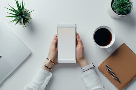 Top view of female hands holding smartphone with blank screen on white office desk. Mock upの素材