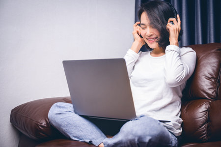 Beautiful asian young woman relaxing with her music playing with laptop on sofa.Work at home and...