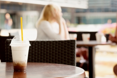 Glass of ice coffee on a table at a cafe with unrecognizable girl out of focus in the backgroundの写真素材