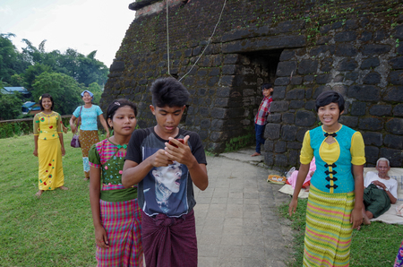 Mrauk-u, Rakhine State / Myanmar - October 18 2016: Traditionally dressed up teenagers and a young man with a mobile phone in contrast to an old beggar in front of an ancient templeのeditorial素材