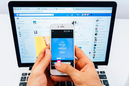 A person is holding a white smartphone with a blue screen in front of an open laptop computer. The laptop screen displays a web page with various elements. The person's hands are visible, with their thumbs positioned over the phone's screen. The setup suggests a comparison or synchronization task between the two devices.