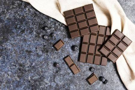 a pile of chocolate squares on a gray marble surface next to a beige cloth with a few chocolate chips scattered around