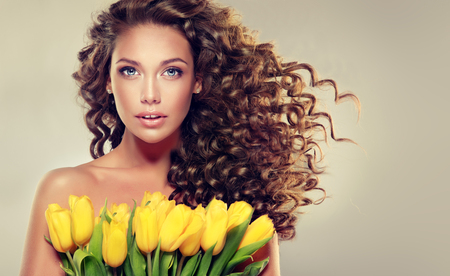 Young brown haired model with dense, curly,voluminous hair is holding bouquet of yellow tulips  on the chest. Simbol of spring, summer and freshness.の写真素材