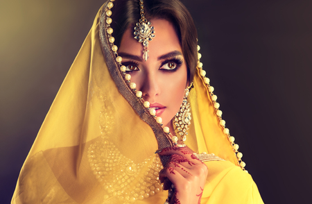 Close up portrait of young indian woman is covering part of the face by yellow cloth with white pearls. Bright make up, long eyelashes and tender look aside.の写真素材