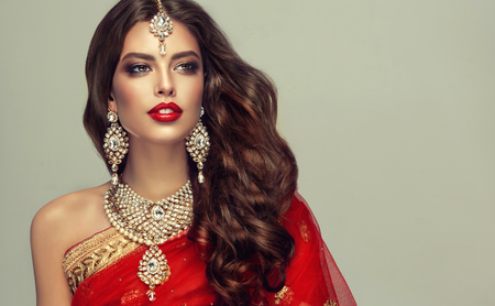 Young attractive woman, covered by traditional indian red shawl (dupatta) and dressed in hand-made kundan style jewelry set. Posh jewelry set is consisting of big earrings, bright necklace and head adornment (tikka). Perfect, dense, wavy, freely flying hair and smoky eyes style makeup.